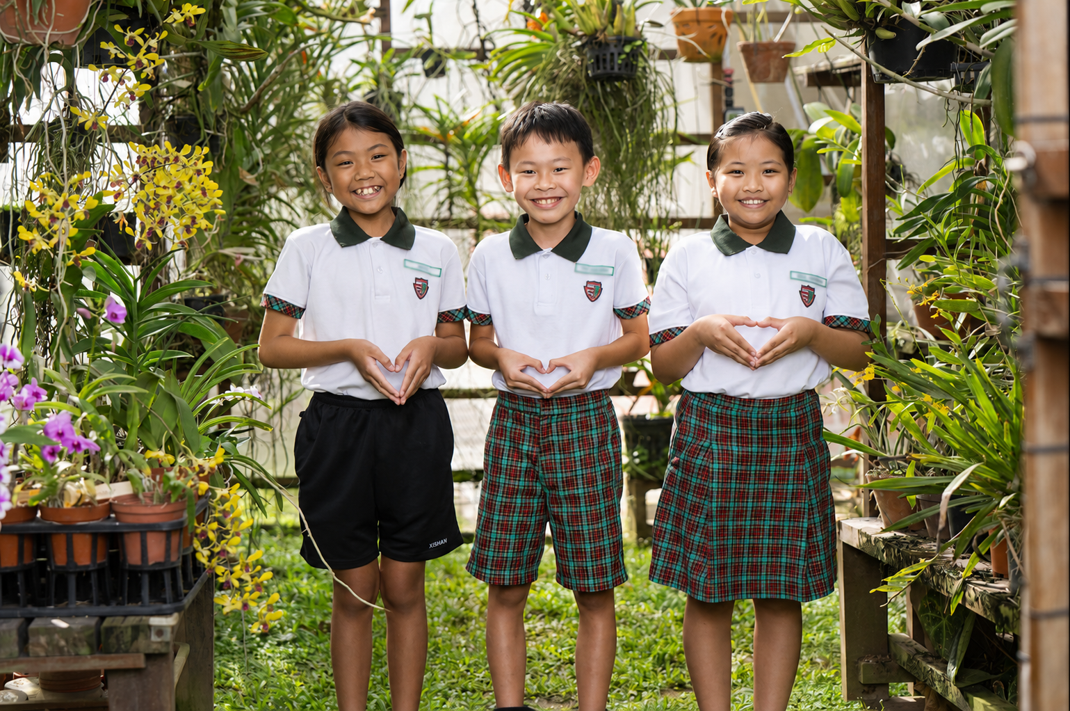 Three smiling students in school uniforms stand together in a lush garden filled with orchids and other plants. Each student forms a heart shape with their hands while facing the camera. They wear white polo shirts with dark green collars and tartan-patterned uniform bottoms.